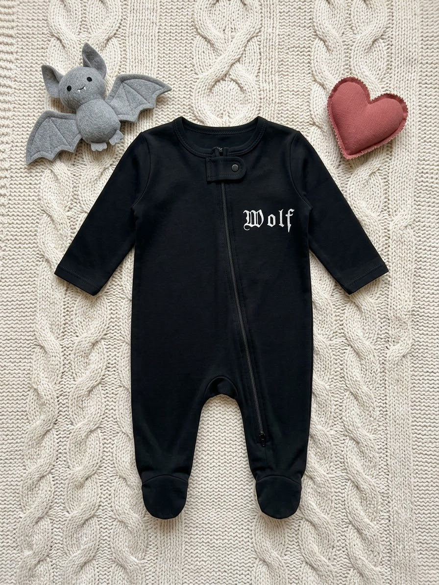 A black baby footed sleeper with long sleeves and 'Wolf' embroidered in white gothic lettering on the chest, laid flat on a cream cable-knit blanket with a gray plush bat and a pink heart prop.