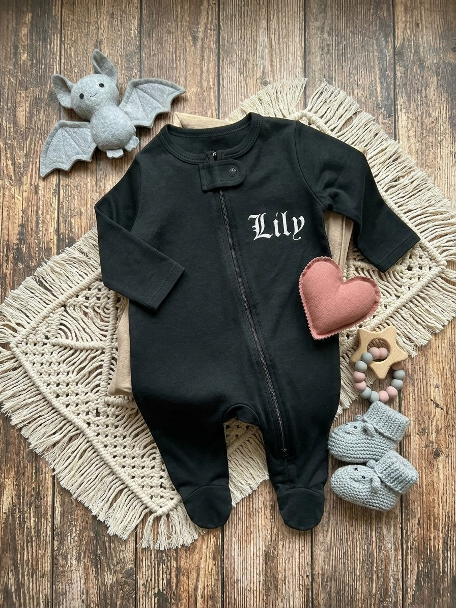 A black baby footed sleeper with 'Lily' embroidered in white Gothic lettering on the chest, styled in a flat lay on a wooden background with a beige macrame mat, a pink heart toy, gray knit booties, and a gray plush bat.