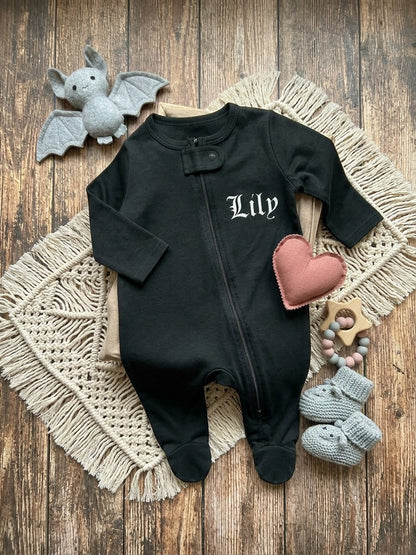 A black baby footed sleeper with 'Lily' embroidered in white Gothic lettering on the chest, styled in a flat lay on a wooden background with a beige macrame mat, a pink heart toy, gray knit booties, and a gray plush bat.
