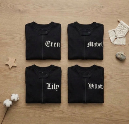 Four folded black baby sweatshirts arranged in a 2x2 grid on a light wood surface. Each sweatshirt has a single white Gothic-style name embroidered on the front.