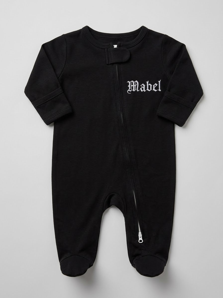 A black baby footed sleeper with a front zipper, featuring the name 'Mabel' embroidered in a white Gothic font on the left chest, laid flat against a light gray background.