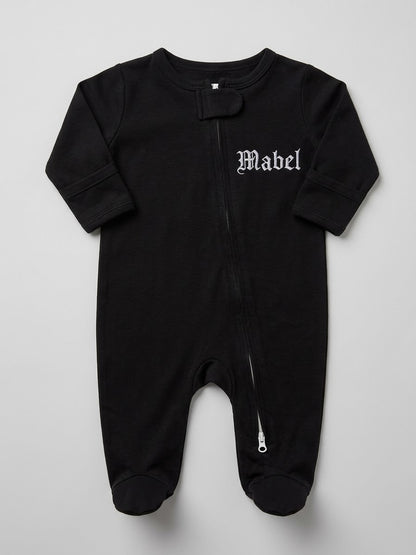 A black baby footed sleeper with a front zipper, featuring the name 'Mabel' embroidered in a white Gothic font on the left chest, laid flat against a light gray background.