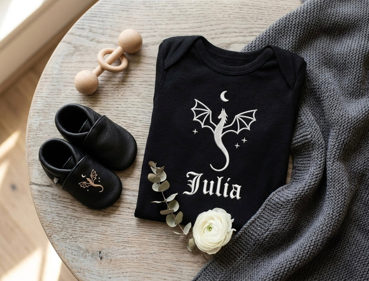 Black baby onesie with dragon design and name 'Julia' on a wooden table with black shoes and a white flower.