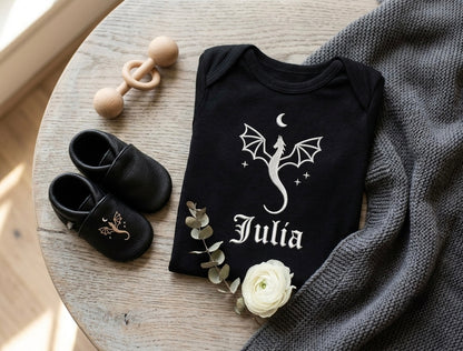 Black baby onesie with dragon design and name 'Julia' on a wooden table with black shoes and a white flower.