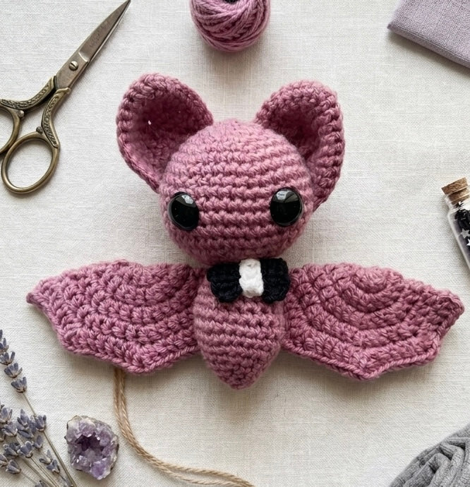 Blush pink handmade crochet bat plush with a black bow tie on a light wood background.