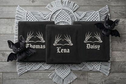 Three black personalized towels laid flat on a white macrame runner over a gray wood plank background. The center towel is folded to show the middle name 'Lena' in white gothic lettering with skeleton hands above, while the left and right towels show the names 'Thomas' and 'Daisy' respectively.