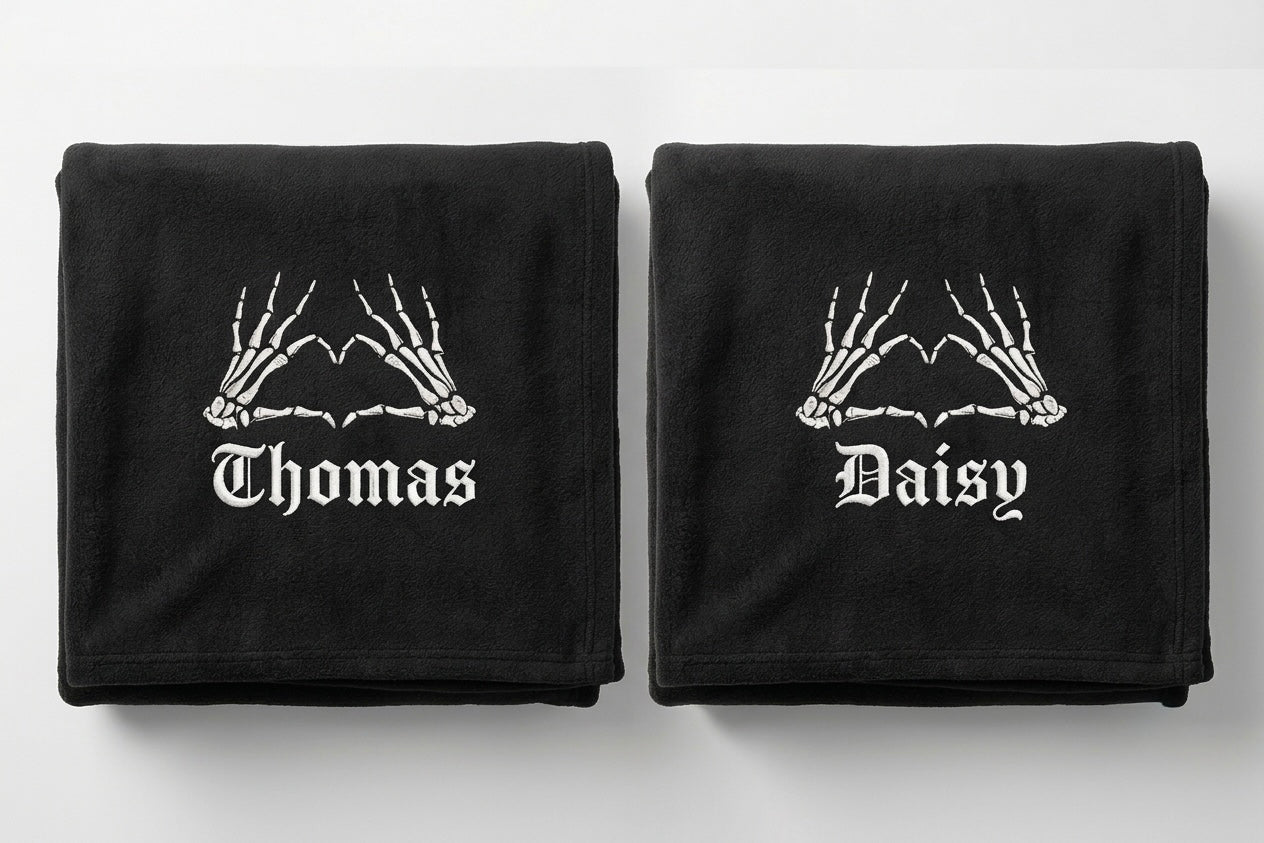 Two black folded towels on a light gray background, each printed with white skeleton hands above the name 'Thomas' on the left towel and 'Daisy' on the right towel.