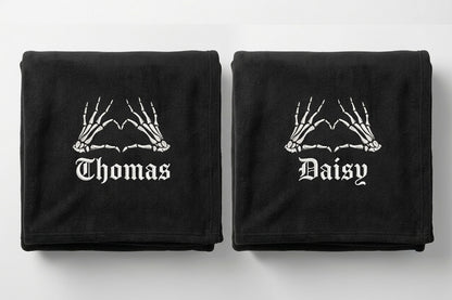 Two black folded towels on a light gray background, each printed with white skeleton hands above the name 'Thomas' on the left towel and 'Daisy' on the right towel.