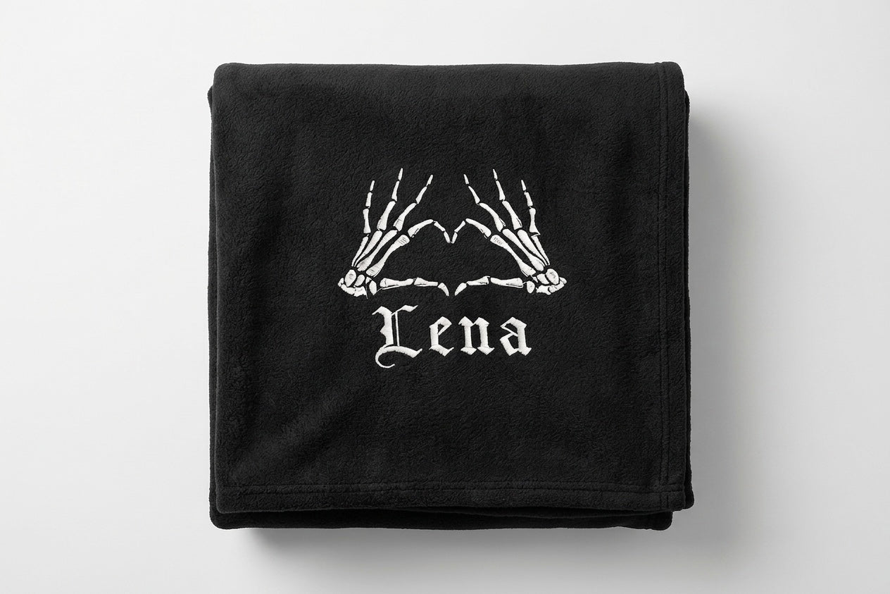 A black folded blanket featuring a white graphic of skeleton hands forming a heart shape above the word 'Lena' in a gothic font, shown on a light gray background.