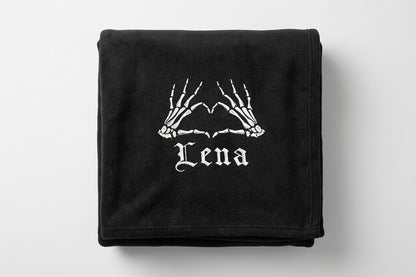 A black folded blanket featuring a white graphic of skeleton hands forming a heart shape above the word 'Lena' in a gothic font, shown on a light gray background.