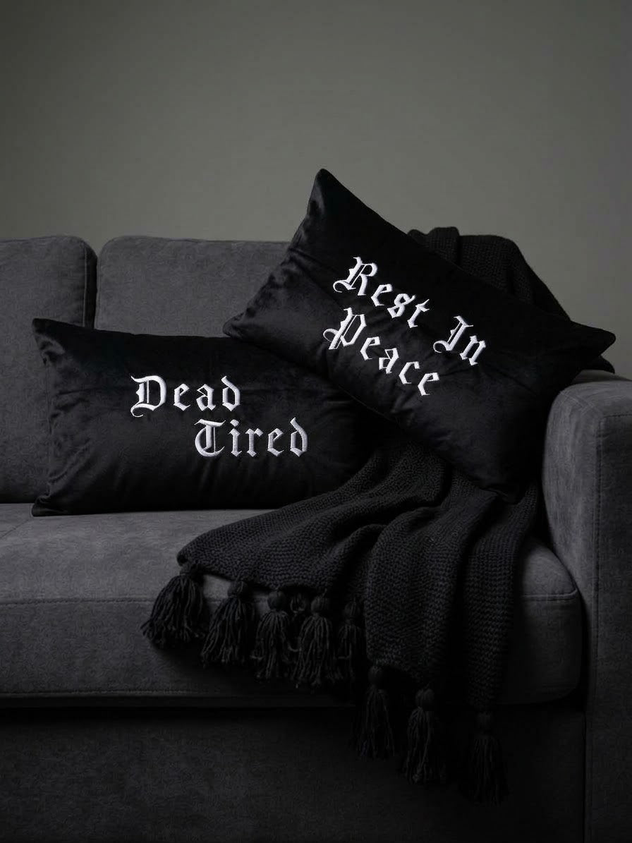Two embroidered gothic black throw pillows with white gothic text reading 'Dead Tired' and 'Rest In Peace' styled on a dark gray sofa with a black fringed throw blanket.