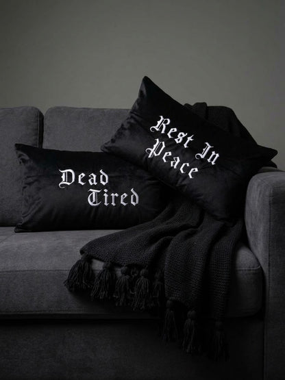 Two embroidered gothic black throw pillows with white gothic text reading 'Dead Tired' and 'Rest In Peace' styled on a dark gray sofa with a black fringed throw blanket.