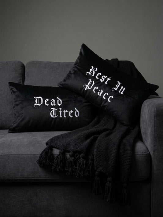 Two embroidered gothic black throw pillows with white gothic text reading 'Dead Tired' and 'Rest In Peace' styled on a dark gray sofa with a black fringed throw blanket.