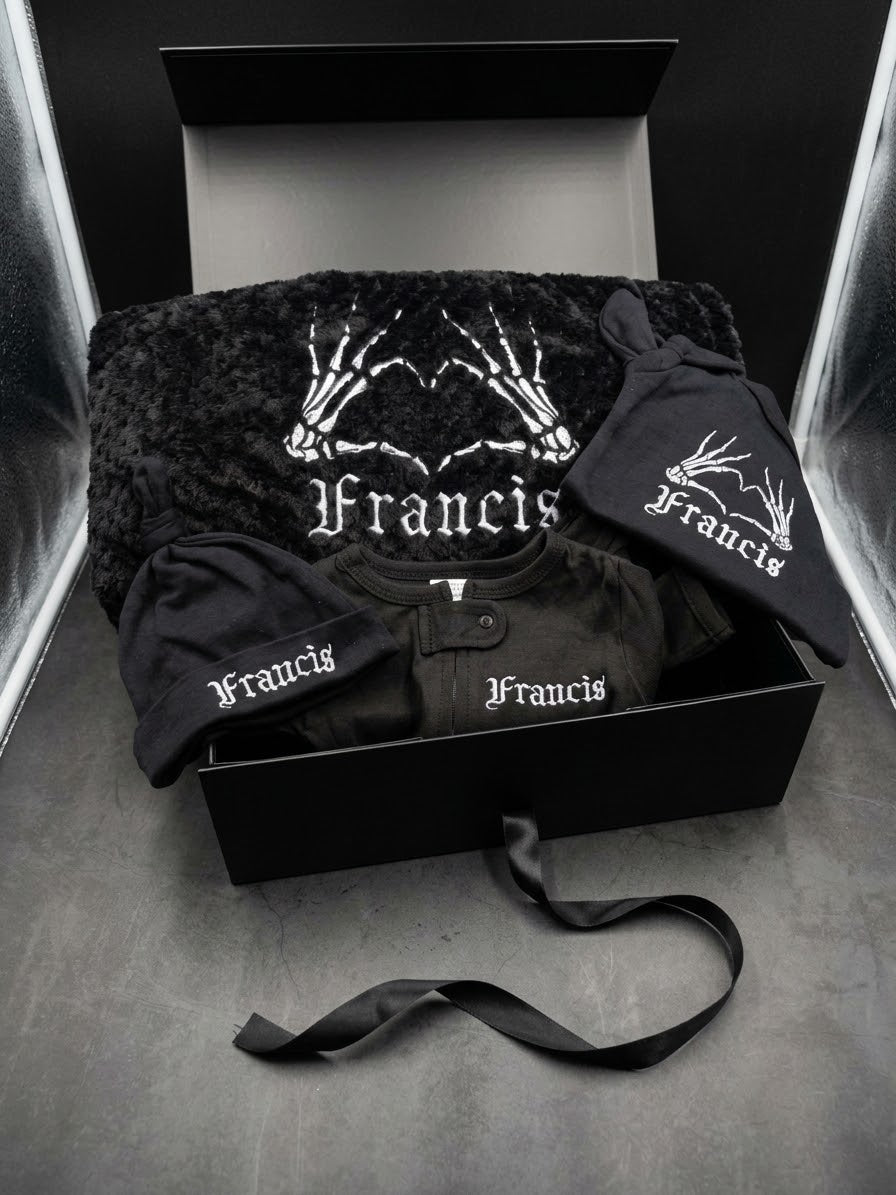 A black baby clothing set from the brand Francis, including a plush blanket, a short-sleeve shirt, and two hats, all featuring white skeleton hand graphics and the brand name 'Francis' in a gothic font, presented in an open black gift box.
