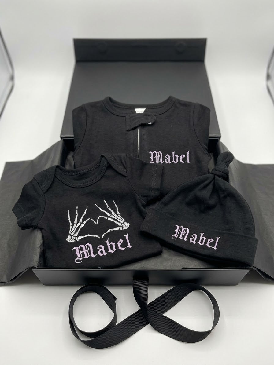 A black personalised baby clothing set displayed inside an open black gift box on a white surface. The set includes a long-sleeve top, a short-sleeve bodysuit, and a knotted beanie, all printed with the name 'Mabel' in white gothic lettering.