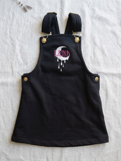 A black pinafore overall-style 
 dress or pinafore with adjustable shoulder straps, laid flat on a white textured fabric background. The front has a white dripping moon graphic with the word 'Lena' in pink above it, and small gold-tone metal rivets and snaps.