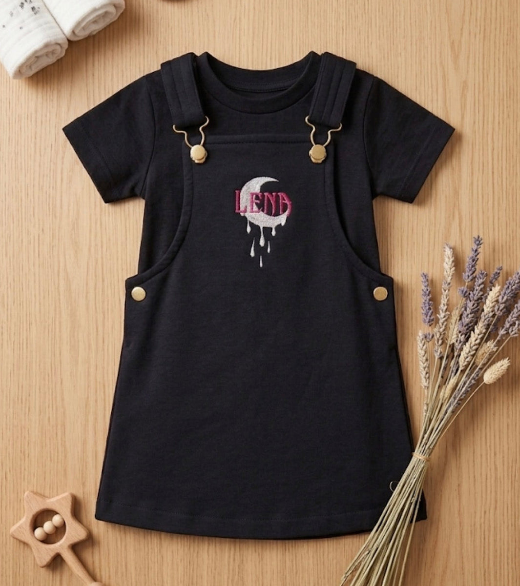 A baby girl's short-sleeve black dress styled as overalls, featuring a front graphic of a dripping heart with the name 'Lena' in pink and white. The dress is displayed in a flat-lay arrangement on a light wood background with baby accessories including a star rattle, lavender, baby shoes, and a rolled blanket.