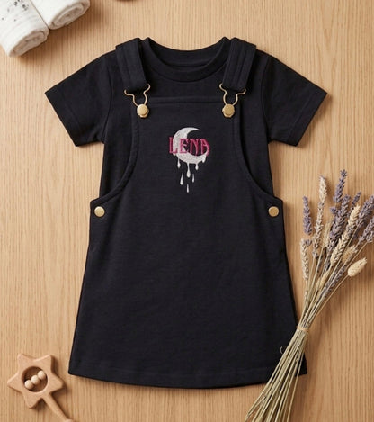A baby girl's short-sleeve black dress styled as overalls, featuring a front graphic of a dripping heart with the name 'Lena' in pink and white. The dress is displayed in a flat-lay arrangement on a light wood background with baby accessories including a star rattle, lavender, baby shoes, and a rolled blanket.