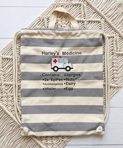 A personalised canvas drawstring bag with gray and cream horizontal stripes, featuring a custom embroidered medical-themed print that reads 'Harley's Medicine' and lists allergies. The bag is styled on a cream macrame wall hanging over white wooden planks.