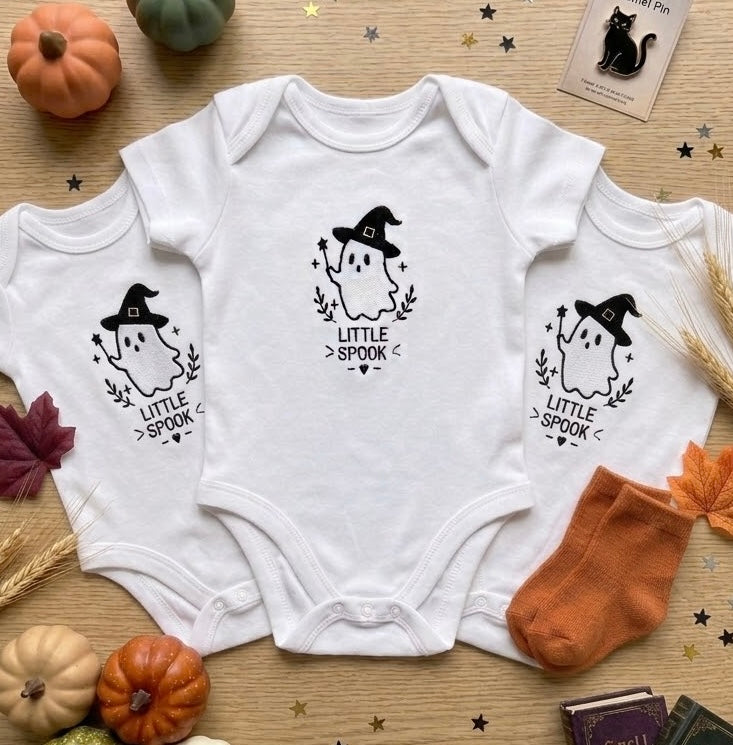 Three white baby bodysuits with a black 'Little Spook' ghost embroidery design are arranged in a flat lay on a wooden surface with autumn props including pumpkins, leaves, a cat card, and socks.
