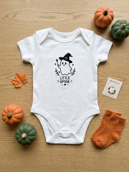 A white short-sleeve baby bodysuit laid flat on a light wood surface, embroidered with a black ghost wearing a witch hat and the words 'LITTLE SPOOK'. Orange and green pumpkins, a small orange leaf, a pair of orange socks, and a small card are arranged around it as seasonal props.