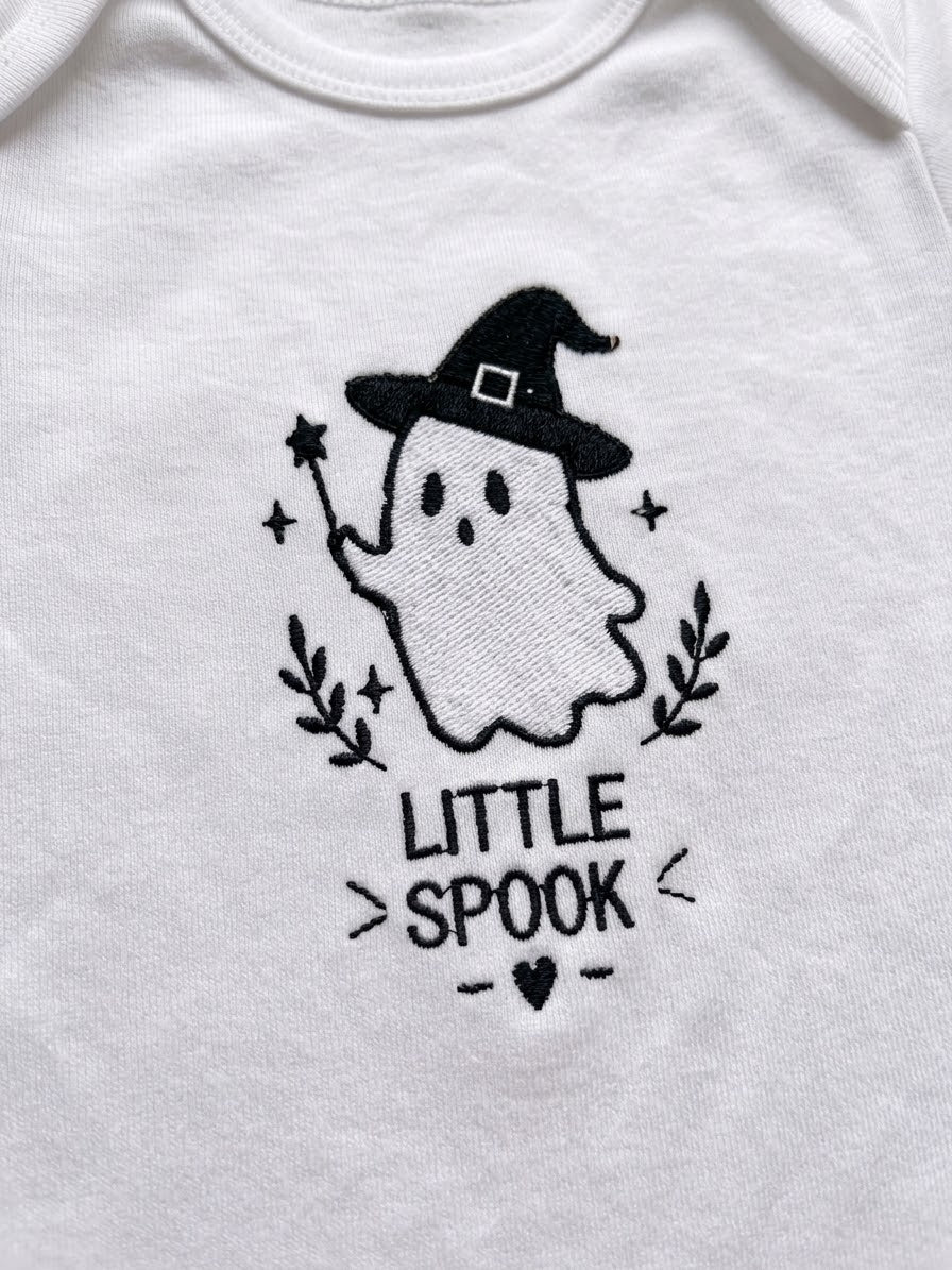 White baby bodysuit featuring a black embroidered graphic of a cute ghost wearing a witch hat with the text 'LITTLE SPOOK' below it.