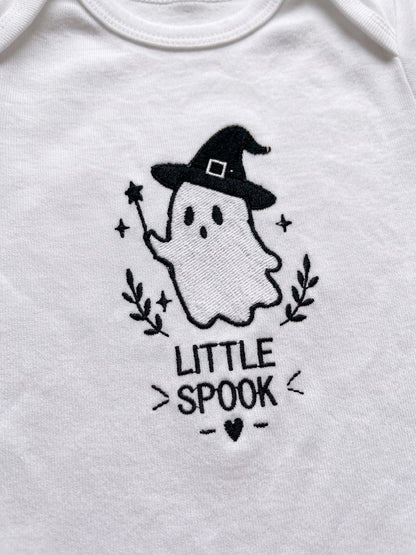 White baby bodysuit featuring a black embroidered graphic of a cute ghost wearing a witch hat with the text 'LITTLE SPOOK' below it.