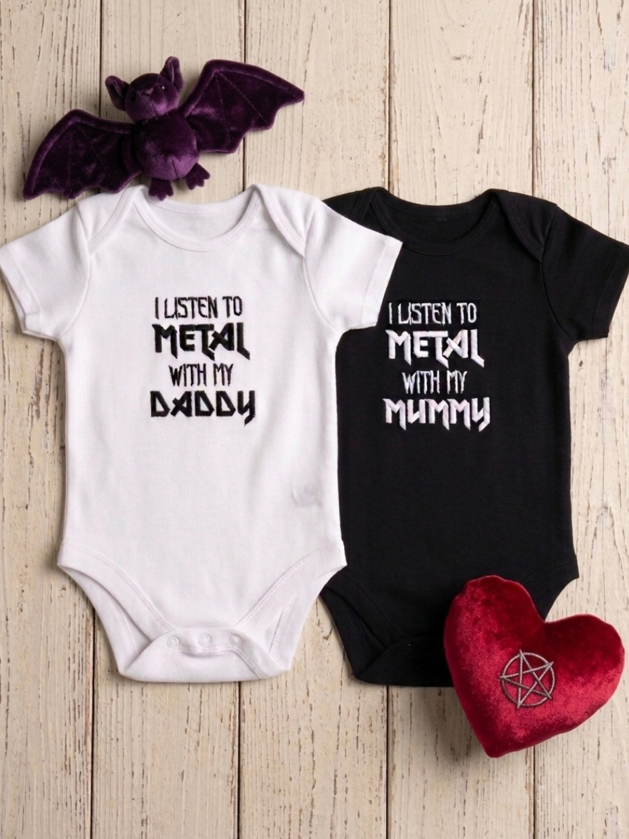 Two baby bodysuits laid flat on a light wood background: one white with black embroidered text reading 'I LISTEN TO METAL WITH MY DADDY' and one black with white text reading 'I LISTEN TO METAL WITH MY MUMMY'. A purple bat plush and a red heart plush with a pentagram symbol are placed around them as props.