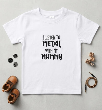 A white kids' short-sleeve t-shirt with embroidered black text reading 'I LISTEN TO METAL WITH MY MUMMY' laid flat on a light gray surface, surrounded by small styled props including brown shoes, skull pins, wooden blocks, and an audio cable.