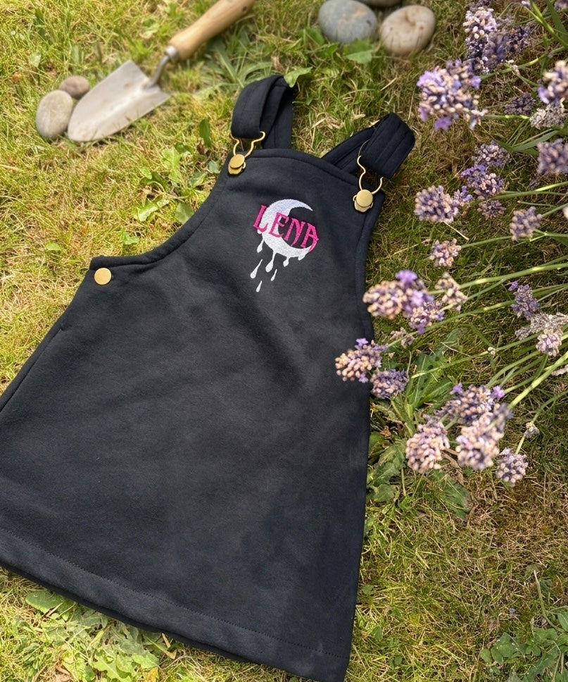 A black dungaree pinafore dress laid flat on grass next to purple flowers, with a small trowel and stones in the background. The apron has a pink and white embroidered logo on the chest.