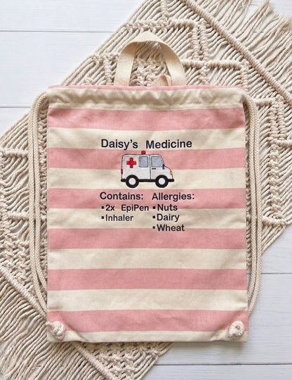 A fabric medicine bag with pink and cream horizontal stripes, featuring an embroidered ambulance graphic and allergy list text reading 'Daisy's Medicine' and 'Contains: Allergies:'. The bag is laid flat on a cream macrame rug over a white wood background.