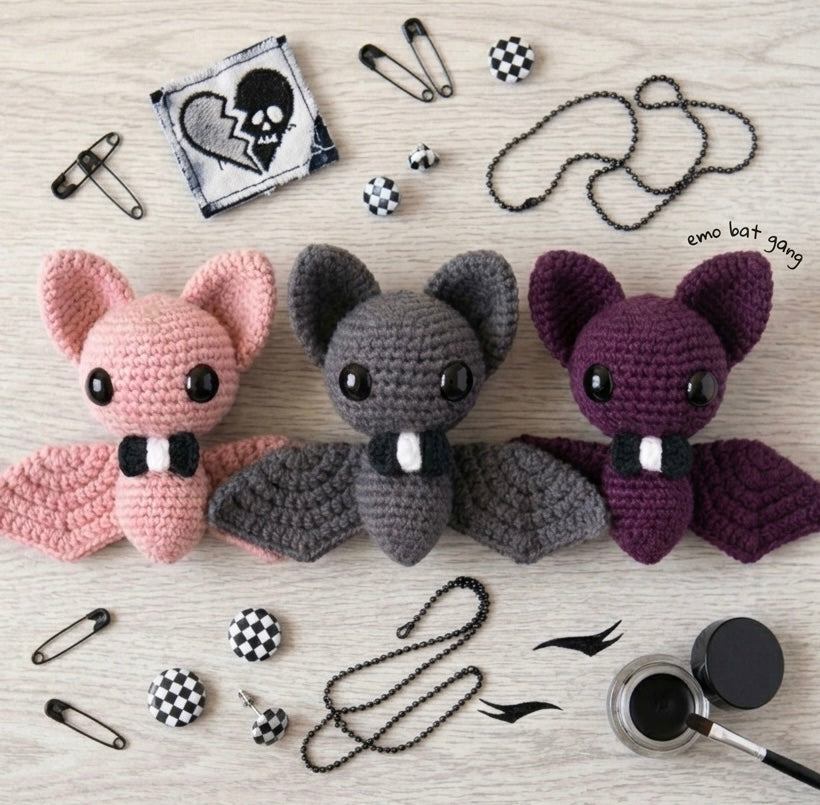 Flat lay photograph of three crochet bat plushies in pink, grey, and purple surrounded by emo accessories 