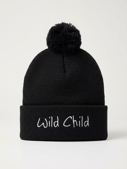 A black knit pom-pom beanie with the words 'Wild Child' embroidered in white script on the front cuff, shown against a plain light background.