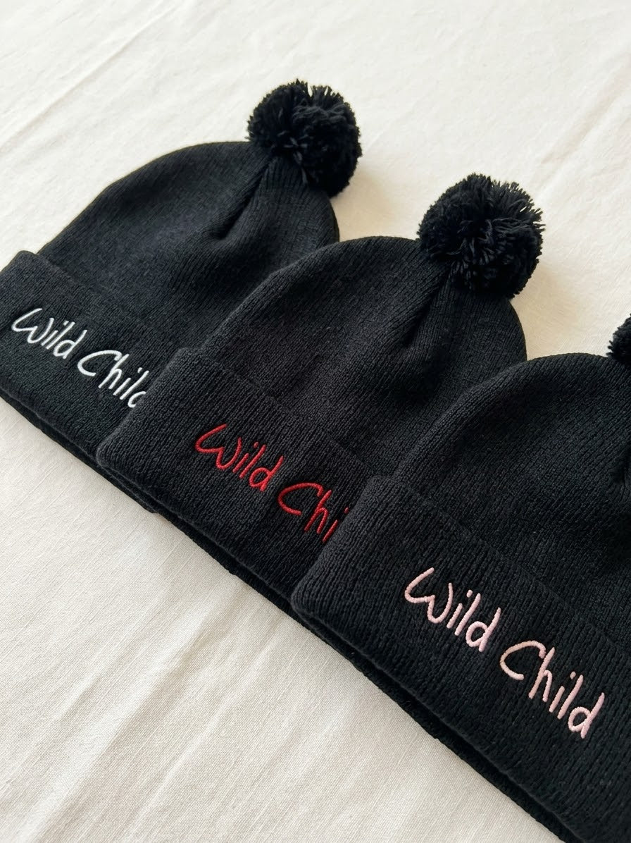 Three black knit pom-pom beanies laid diagonally on a white fabric background, each with the words 'Wild Child' embroidered on the cuff in different colors: white, red, and light pink.