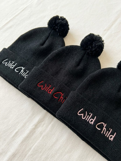 Three black knit pom-pom beanies laid diagonally on a white fabric background, each with the words 'Wild Child' embroidered on the cuff in different colors: white, red, and light pink.