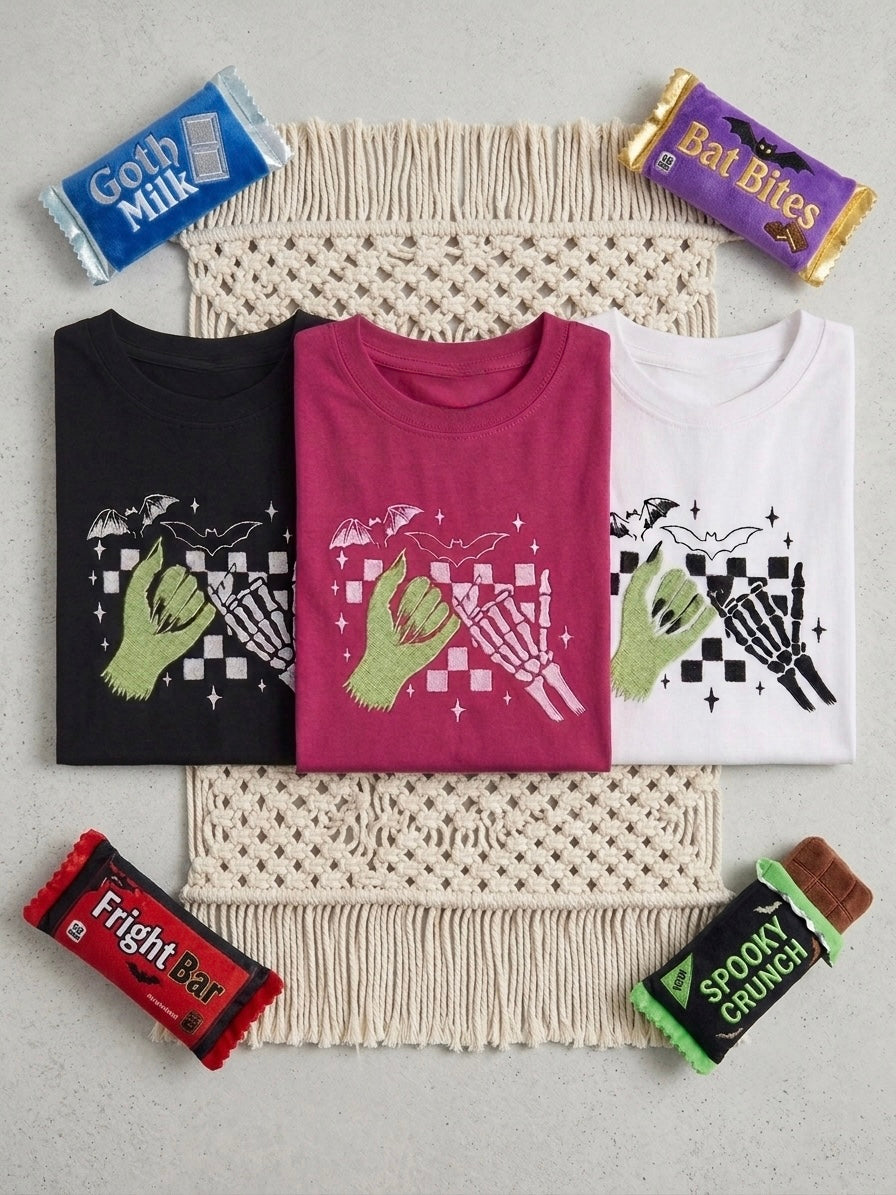 Three folded embroidered gothic alternative graphic T-shirts in black, magenta, and white are arranged on a cream woven mat with macrame fringe, surrounded by candy-themed props including Goth Milk, Bat Bites, Fright Bar, and Spooktacular Crunch.