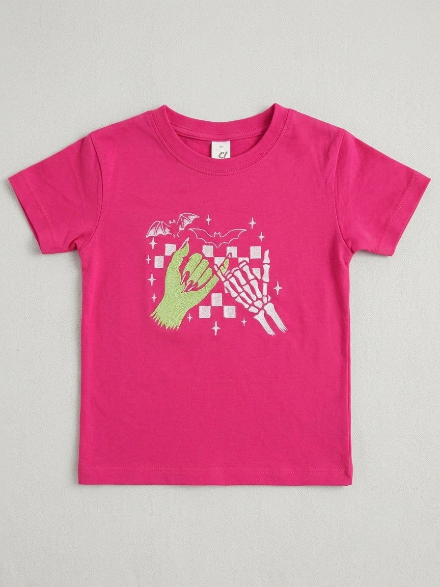 A bright pink short-sleeve t-shirt with a graphic embroidered gothic altnerative zombie on the front showing a green hand, a white skeleton hand, and a pink-and-white checkerboard pattern with stars, laid flat on a light gray background.