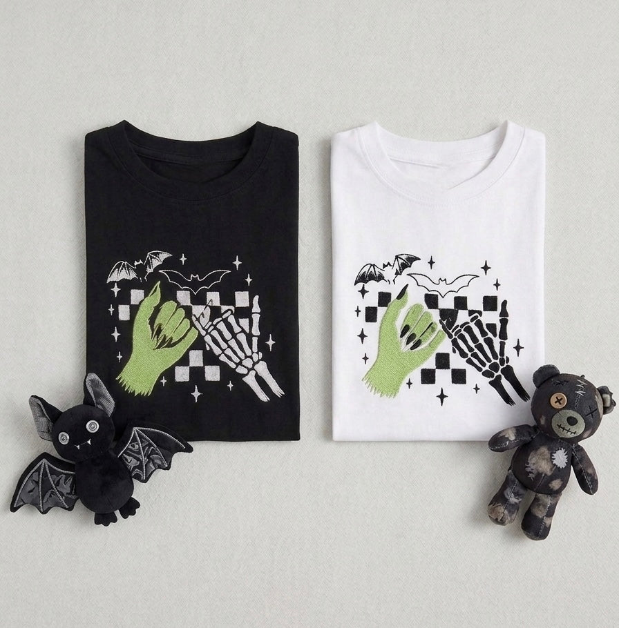 Two folded graphic embroidered alternative gothic zombie T-shirts displayed side by side on a light gray background, with a small black bat plush on the left and a small dark teddy bear plush on the right. The left shirt is black and the right shirt is white, both featuring a gothic graphic of a green hand, skeleton hand, checkered pattern, bats, and stars.
