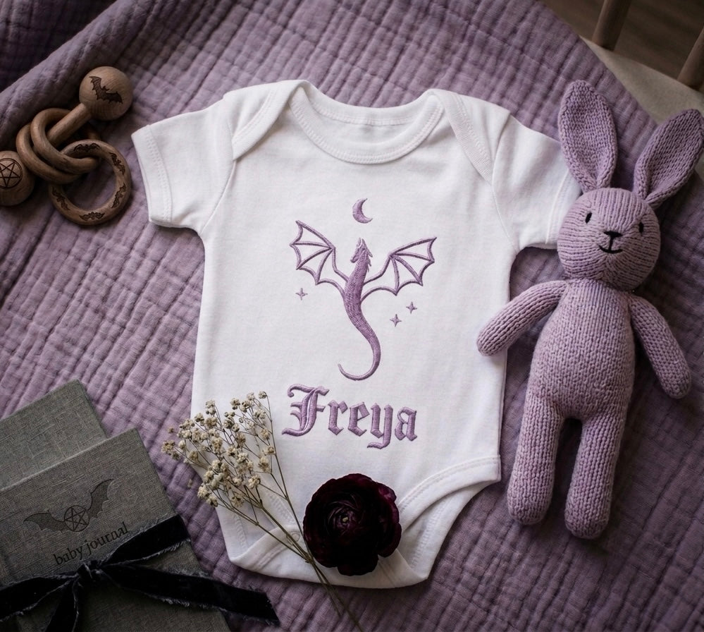 White baby onesie with purple dragon design and 'Freya' text on a purple textured surface with toys and flowers.