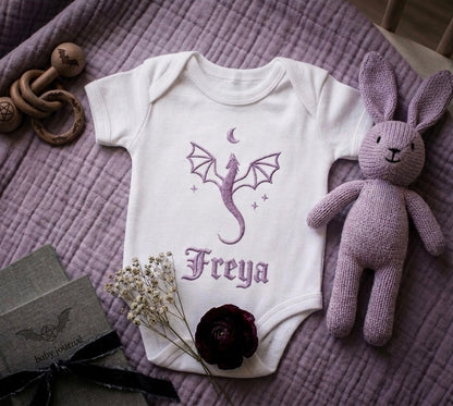 White baby onesie with purple dragon design and 'Freya' text on a purple textured surface with toys and flowers.